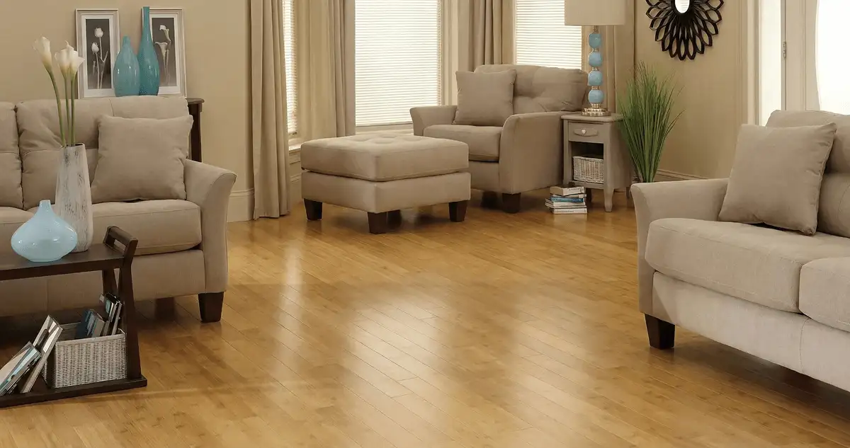 A living room with professionally installed bamboo flooring, beige sofas, a matching ottoman, a side table with a blue lamp, vases, framed pictures, and large windows with blinds and curtains in Rockford, IL